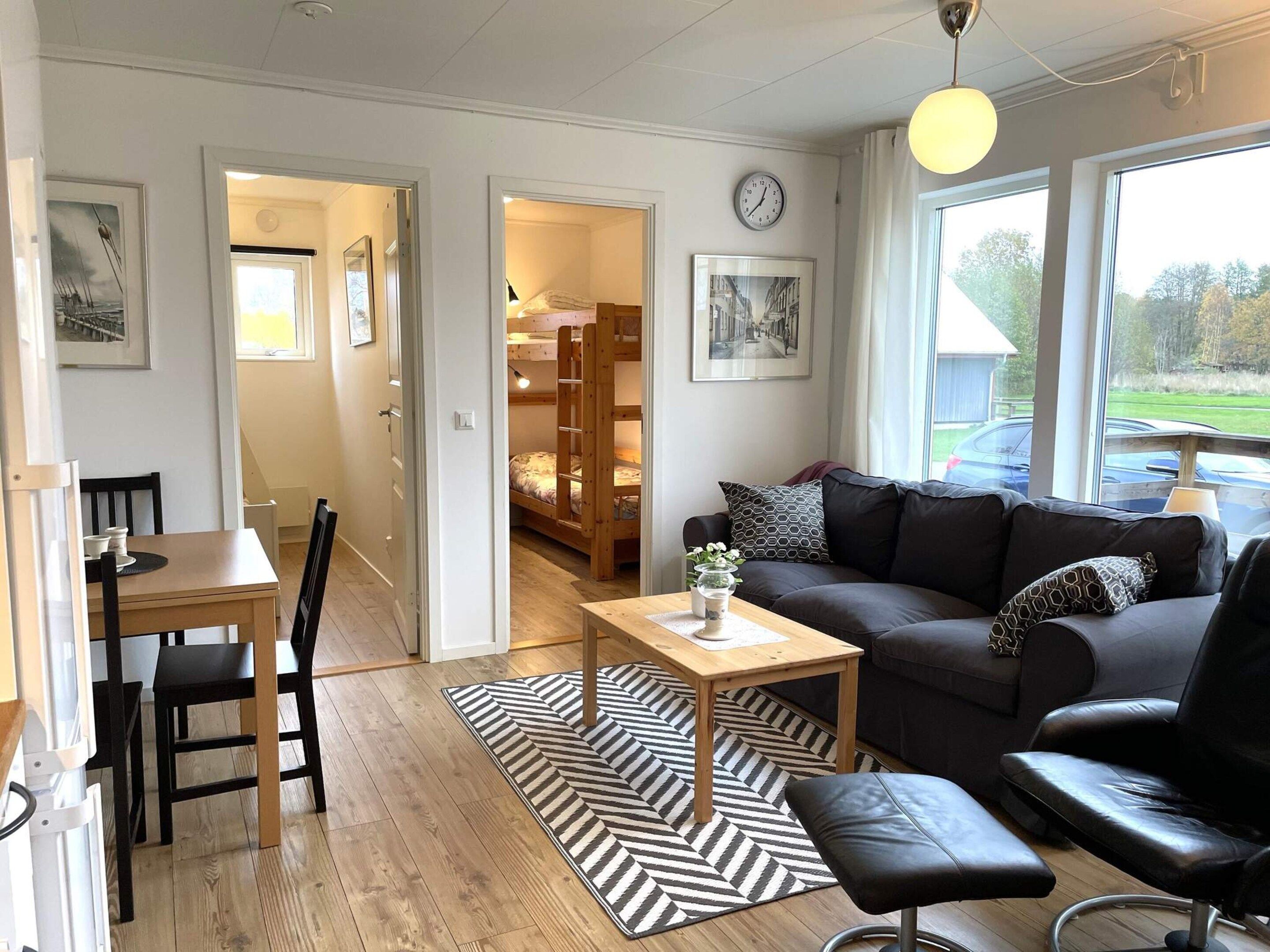 A modern living room with a sofa, a dining table, and a black leather chair. There is a wooden floor, a black and white rug, and a window with a view of the outdoors.