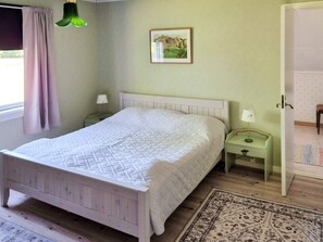 Un dormitorio con cama, mesitas de noche, una lámpara, un cuadro en la pared y una puerta que da a otra habitación.