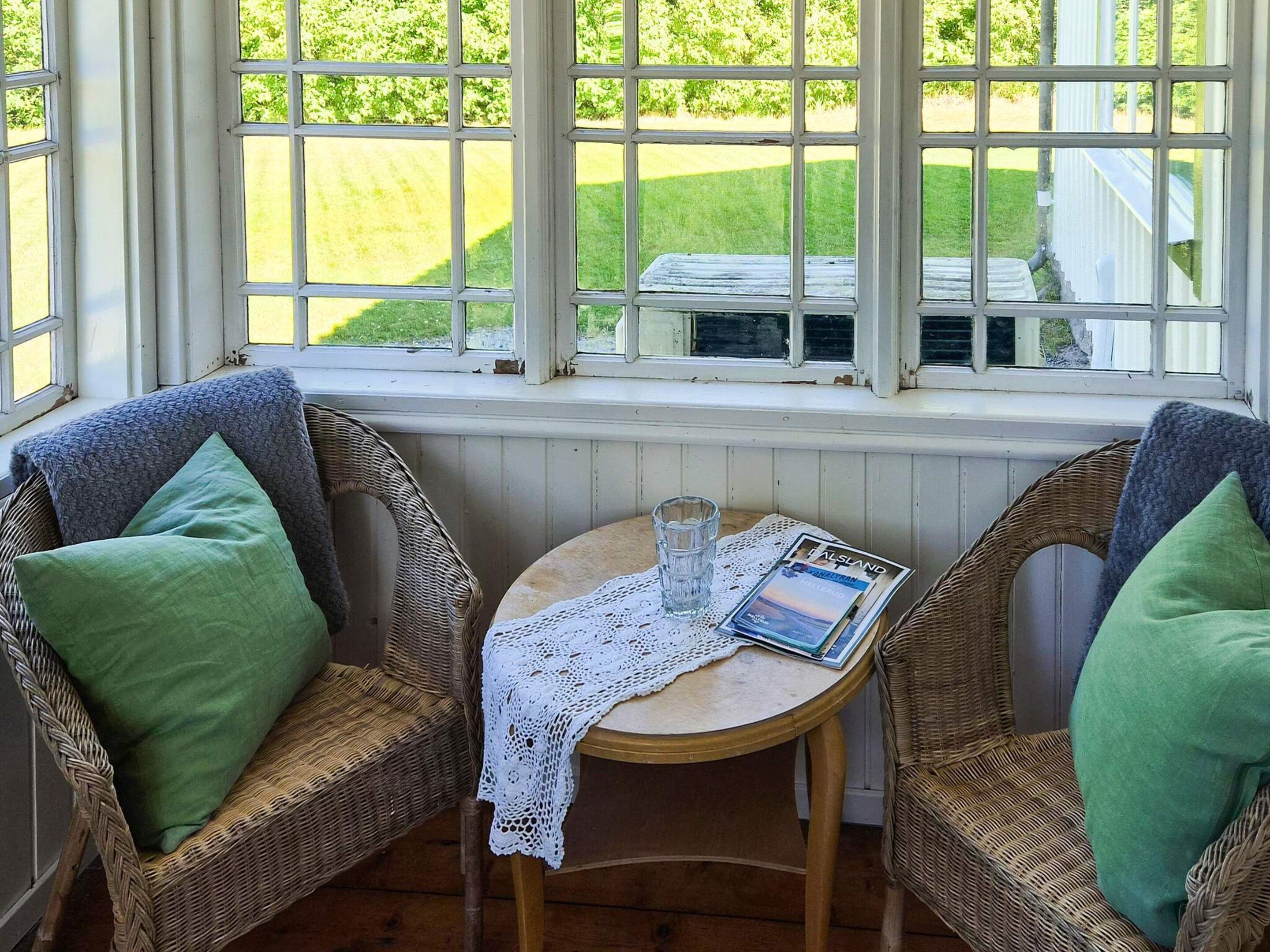 A room with two wicker chairs, a small round table with a glass and a magazine, and a view of a green landscape through large windows.