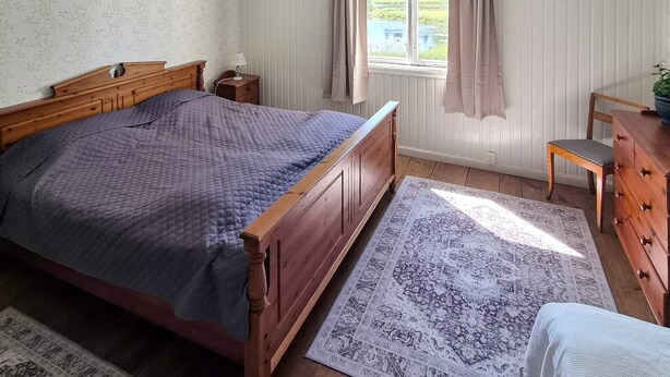 Et soveværelse med en træseng, et natbord, en kommode, et vindue med gardiner og udsigt til grønne områder.