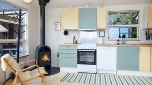 Una cocina moderna con estufa de leña, zona de comedor con una silla y vistas a la vegetación a través del ventanal.