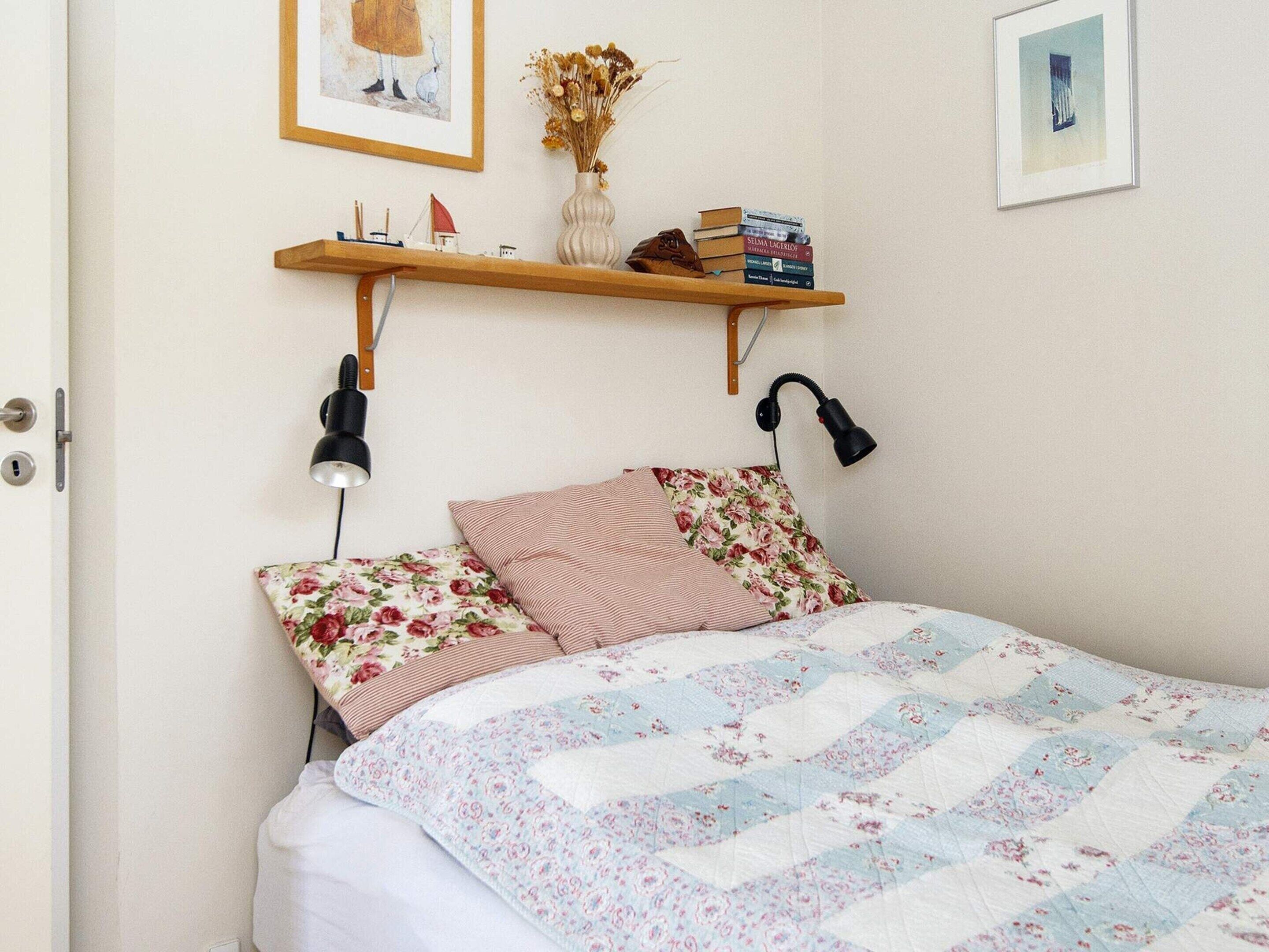 A bedroom with a bed, a nightstand, a shelf with books and a vase, and a door.