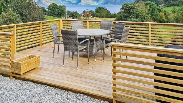 Una terraza de madera con una mesa redonda y sillas de mimbre, con vistas a un lago y montañas.