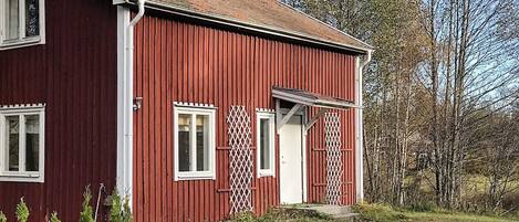 Ett rött trähus med en vit dörr och vita fönster, omgivet av träd och gräs.