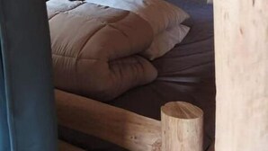 A wooden bunk bed with a single mattress and pillows, a wooden headboard, and a small wooden stool.