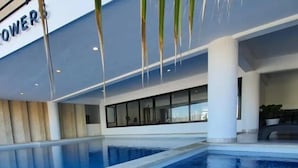 Piscina interior moderna con agua azul cristalina, rodeada por una zona de baldosas blancas y grises, y un ventanal que proporciona luz natural.