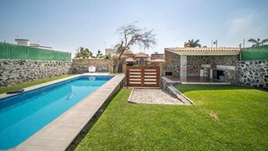 A swimming pool with a stone wall and a wooden gate, surrounded by a well-maintained lawn and a stone building with a tiled roof.