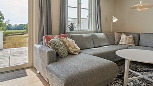 Ett modernt vardagsrum med en grå hörnsoffa, ett runt, vitt soffbord och ett stort fönster med gardiner.