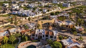 An aerial view of a coastal residential area with modern houses, a central building with a curved facade, and a pool area. - Casas TZL (Cabo San Lucas)