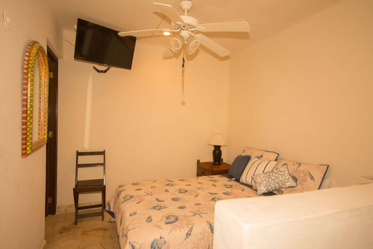 A bedroom with a bed, a chair, a nightstand with a lamp, a ceiling fan, and a TV mounted on the wall.