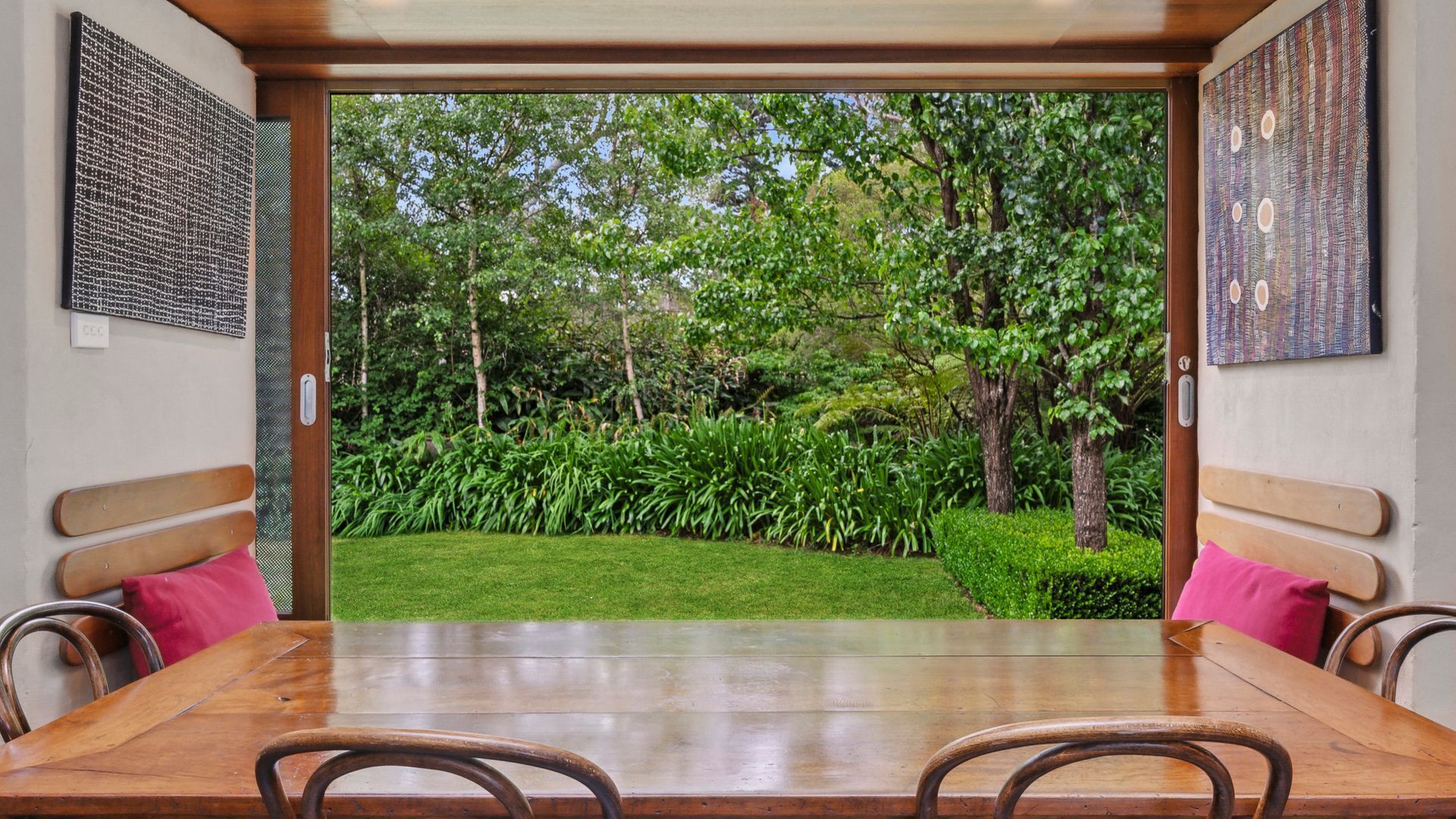 A dining area with a wooden table and chairs, a bench, and a view of a lush garden through an open door.