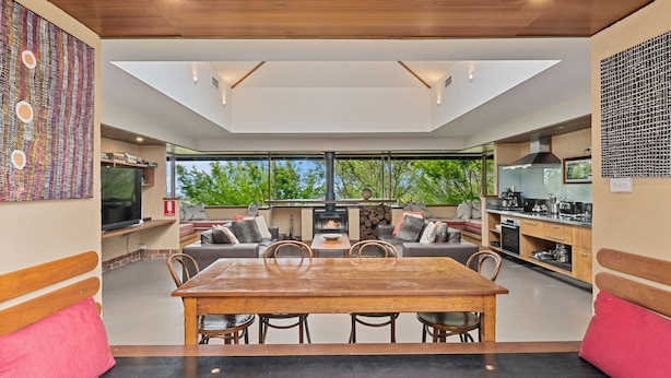 A modern kitchen with a central island, a dining area with a wooden table and chairs, and a large window overlooking greenery. - Summer House - 2 Bedroom House (Wentworth Falls)