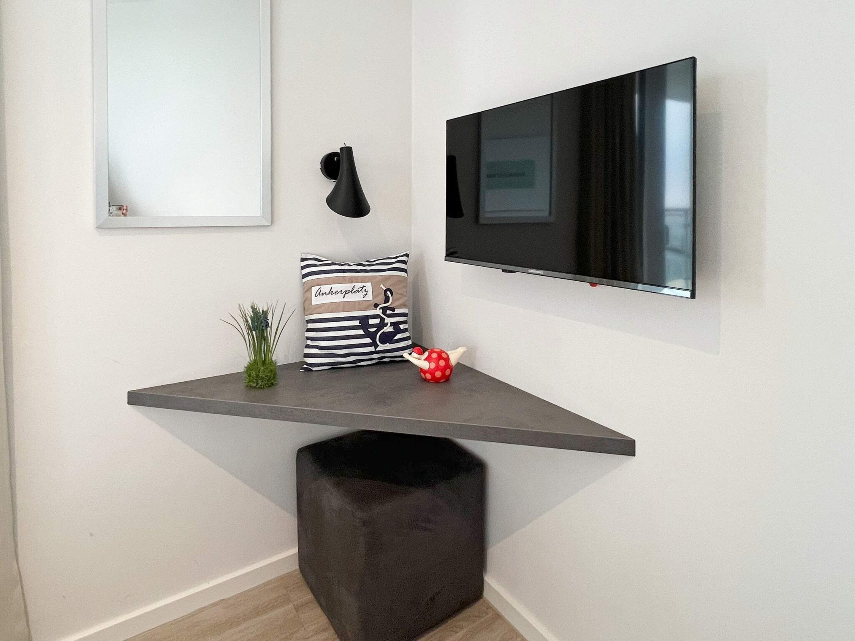 Un espace de vie moderne avec un téléviseur fixé au mur, un luminaire suspendu noir, un coussin à rayures, une petite plante et un jouet rouge posé sur une console sombre.