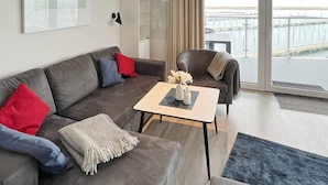 Un salon moderne avec un canapé gris, une petite table et une vue sur un quai, visible à travers de grandes fenêtres.