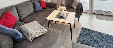 A modern living room with a grey sofa, a small table, and a view of a dock through large windows.