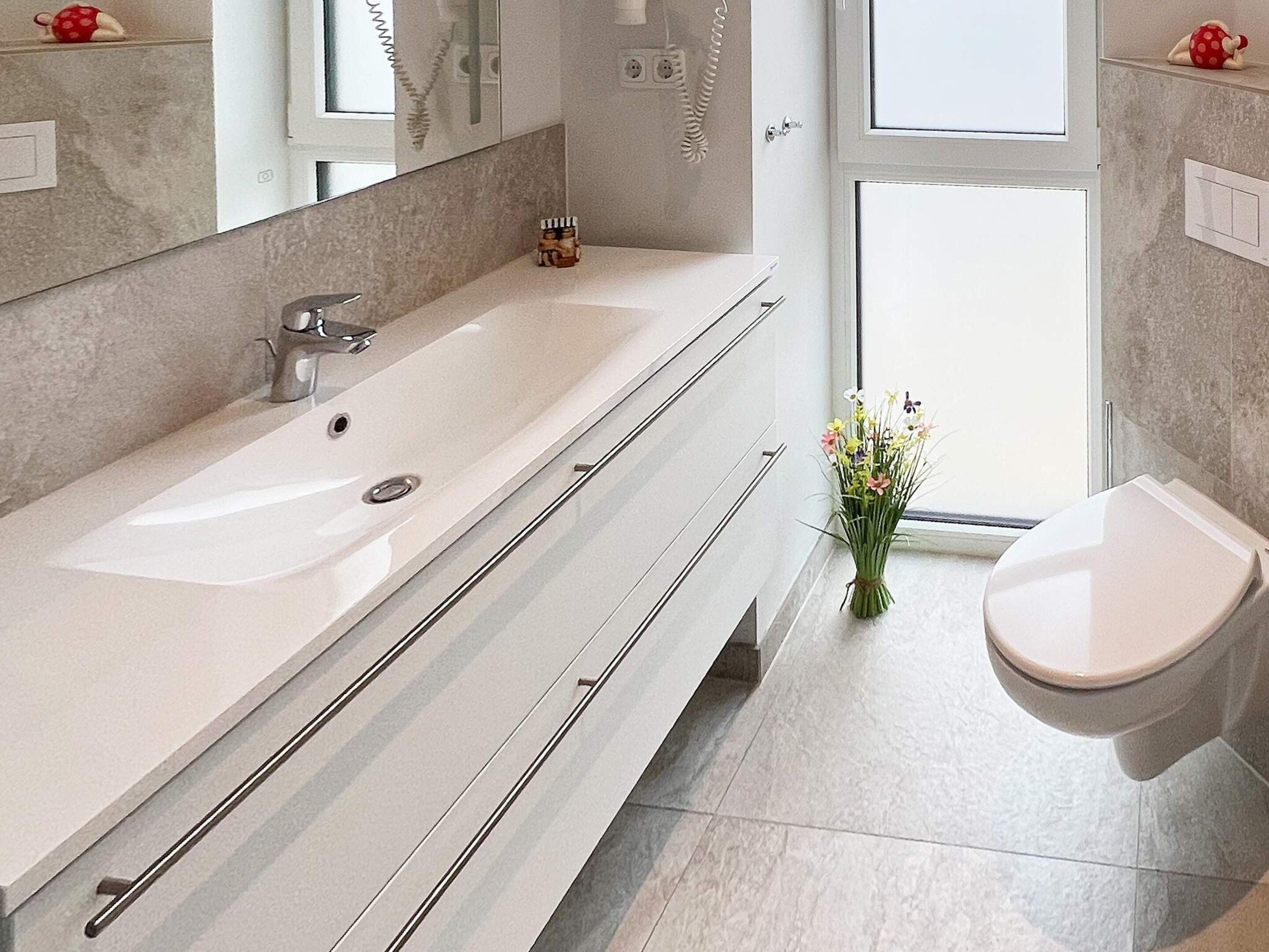 A modern bathroom with a long white sink, a toilet, and a shower area.