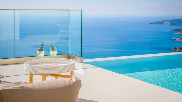 Una piscina infinita moderna con vistas claras al océano y un balcón acristalado con una mesa redonda y dos vasos.