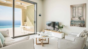 Una sala de estar moderna con un gran ventanal corredizo que da a un balcón con vistas al océano. Hay un televisor de pantalla plana sobre una consola blanca, una mesa de centro con un libro y un área de estar acogedora con sofás.