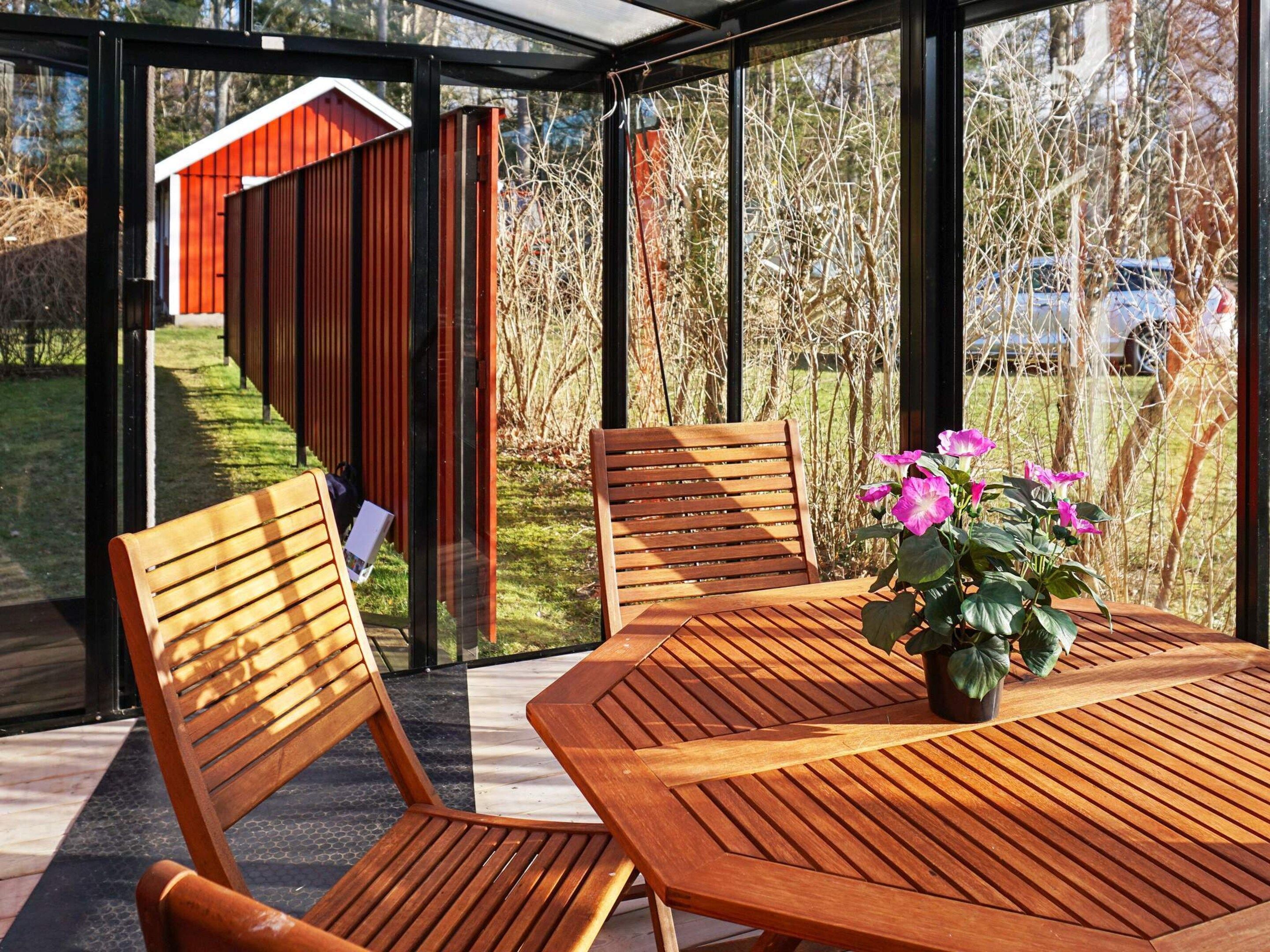 A wooden table with chairs and a potted plant in a sunlit outdoor area with a view of a red building and trees.