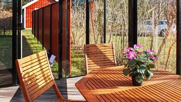 A wooden table with chairs and a potted plant in a sunlit outdoor area with a view of a red building and trees.