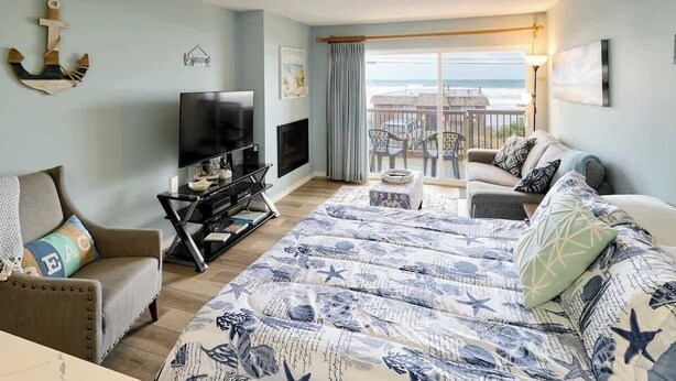 A bedroom with a large bed, a TV, a fireplace, and a balcony with a view of the sea. - Cozy Ocean View Condo - Ground Floor - Fireplace! (Lincoln City)