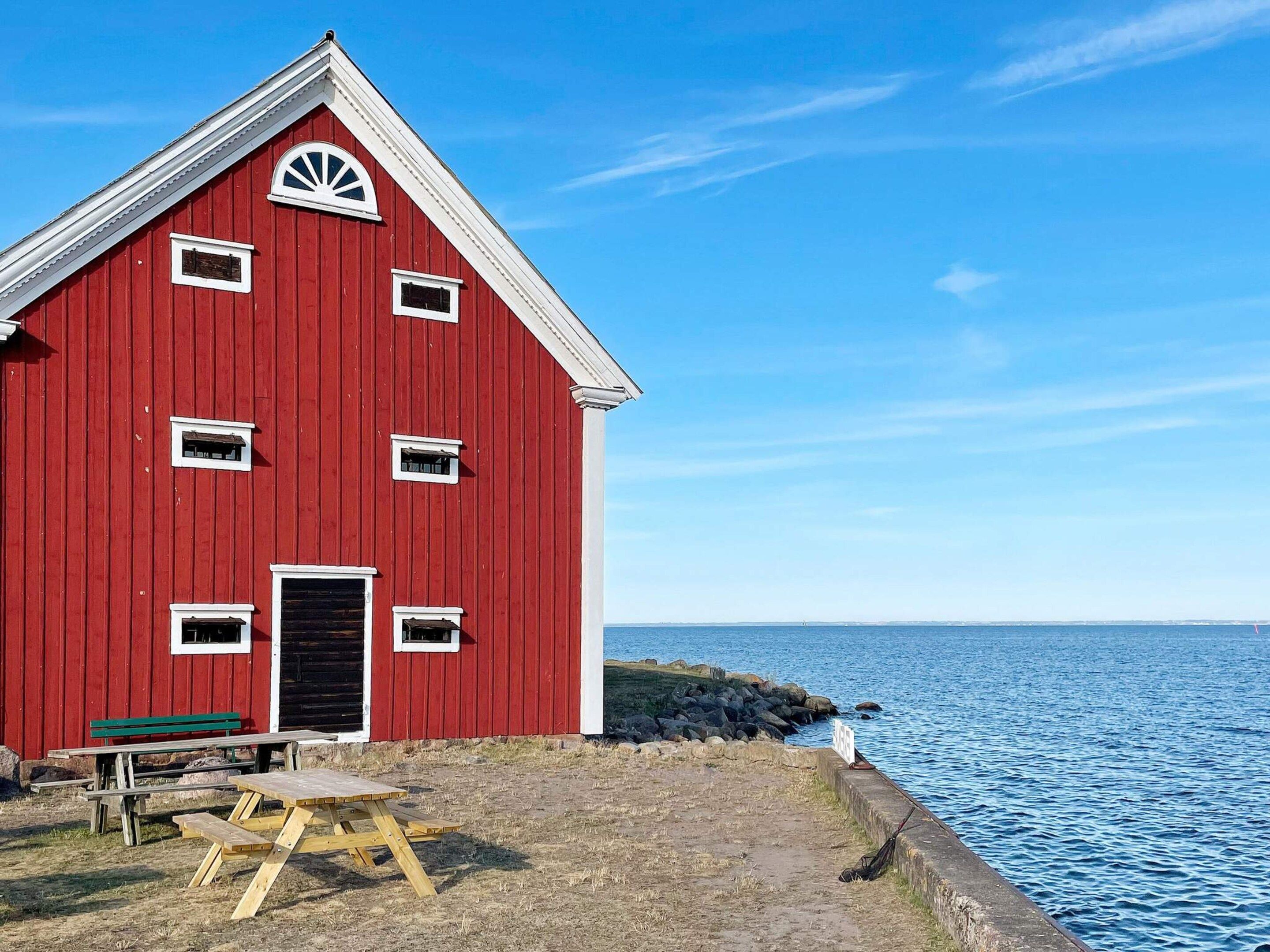 Ein rotes Bauernhaus mit weißer Tür und Fenstern, einem Picknicktisch und Blick auf das Wasser.