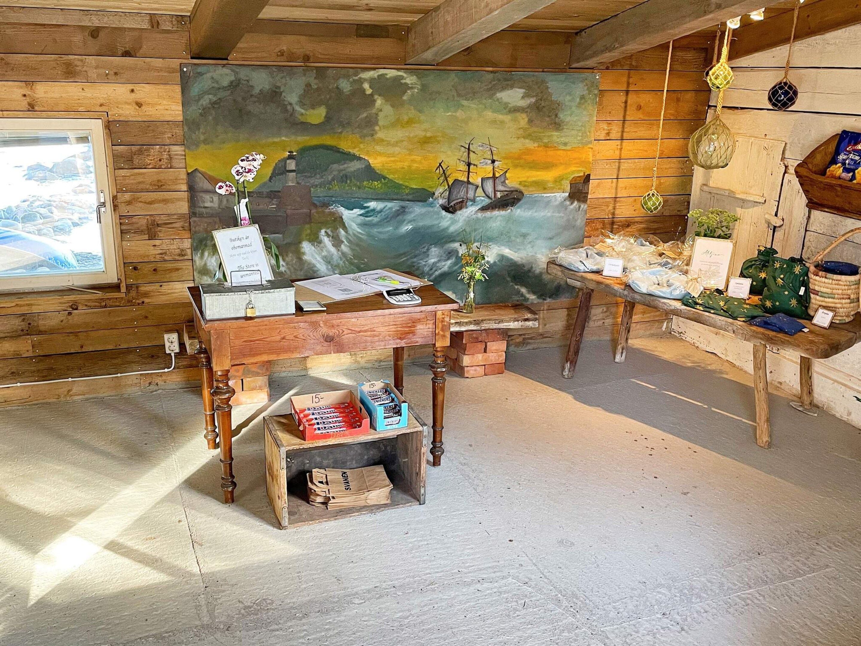 A rustic room with a wooden desk, a painting of a seascape, and a window overlooking a rocky landscape.