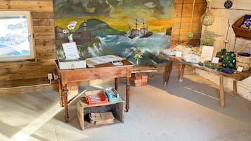 A rustic room with a wooden desk, a painting of a seascape, and a window overlooking a rocky landscape.