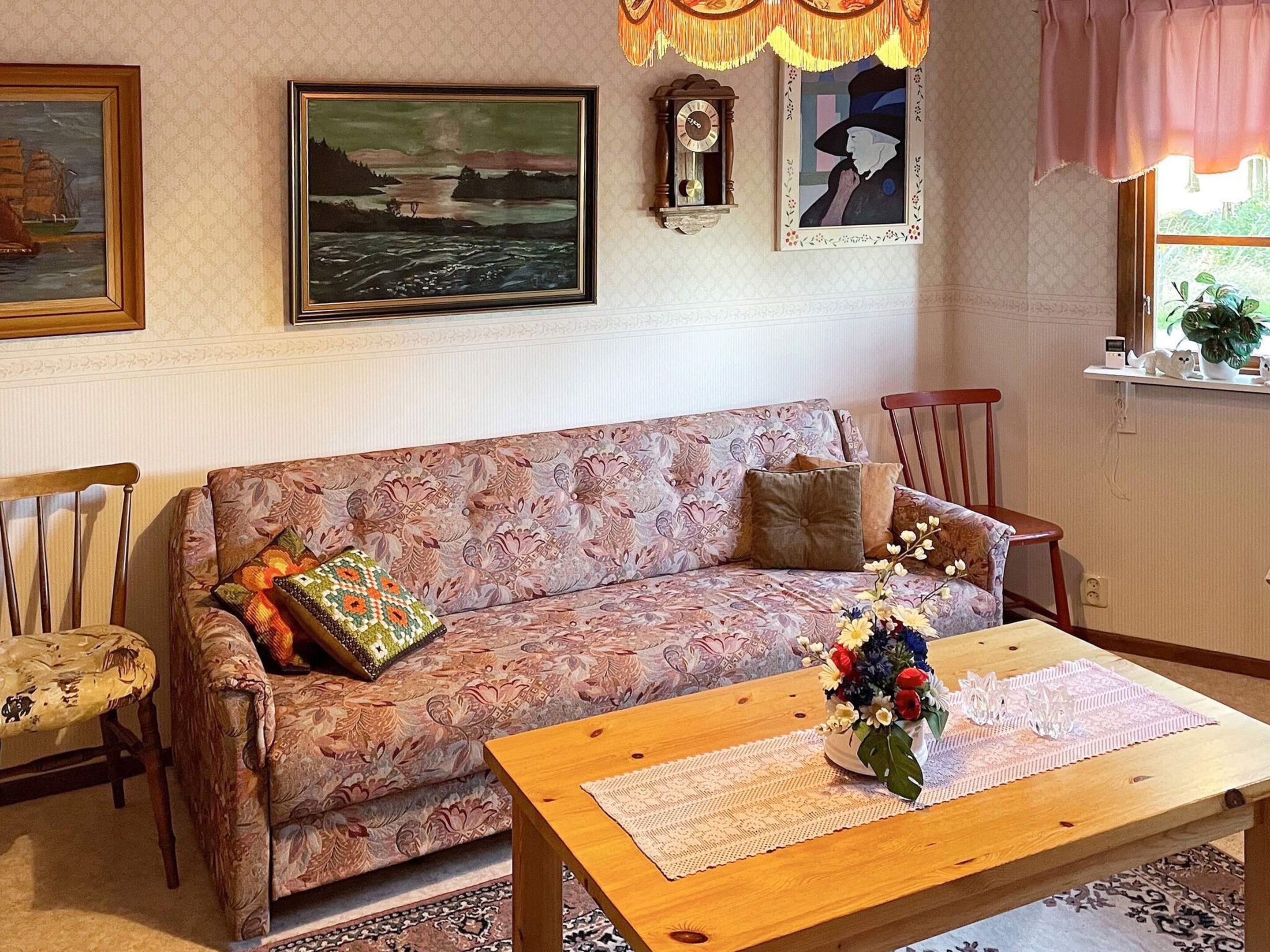 Una sala de estar con un sofá estampado, una mesa de centro de madera con un florero, un reloj y cuadros en la pared.