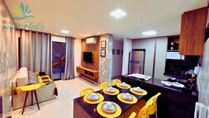 A modern living room with a sofa, a dining table set for six, and a kitchen area with a refrigerator and microwave. - 1. Flat Premium Porto de Galinhas (Ipojuca)