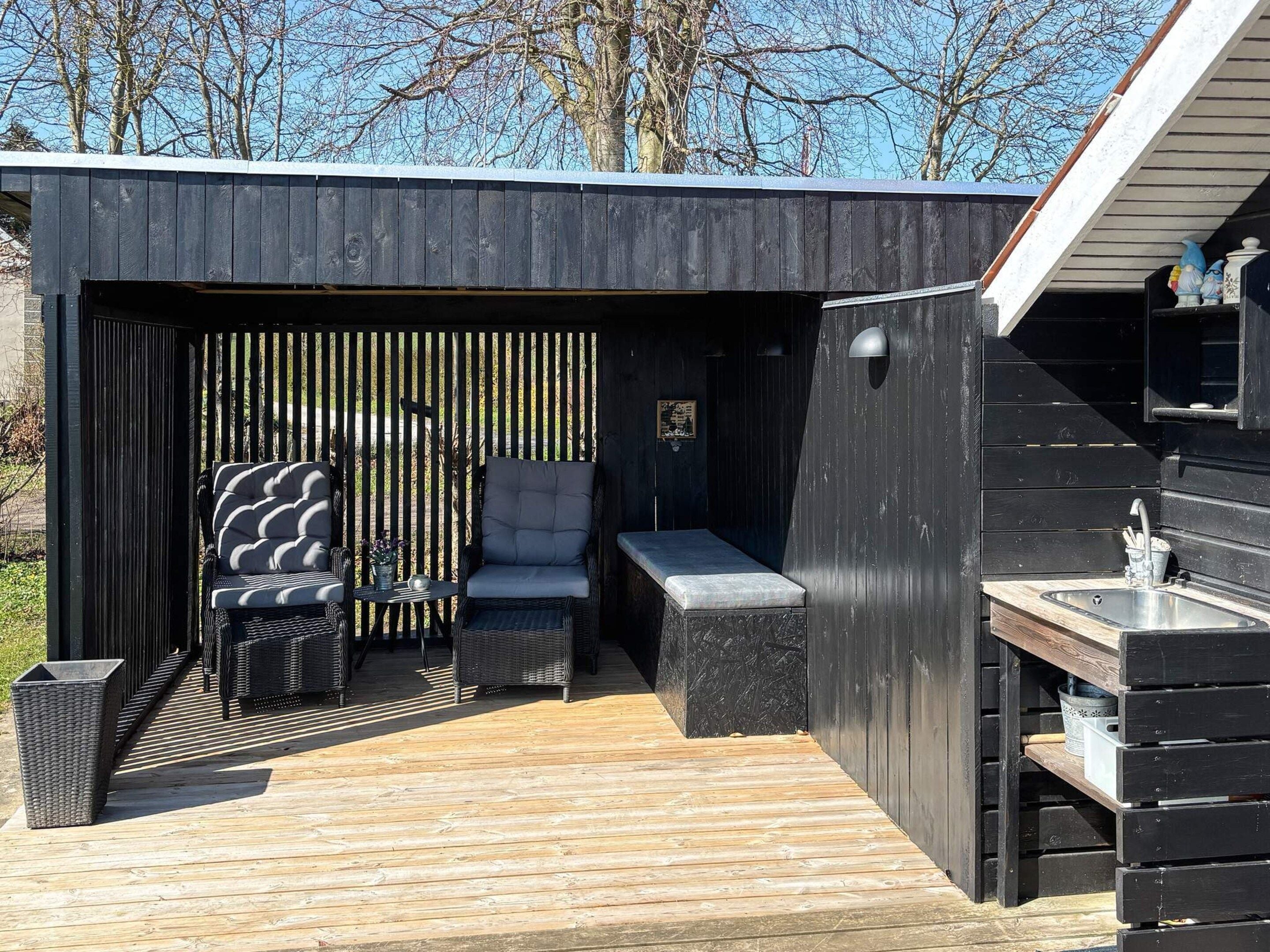 A modern outdoor seating area with a bench, chairs, and a table, surrounded by a wooden deck and a black wall with vertical slats.