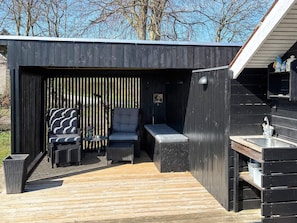 Un espace de détente extérieur moderne, doté d’un banc, de chaises et d’une table, entouré d’une terrasse en bois et d’un mur noir à lattes verticales.