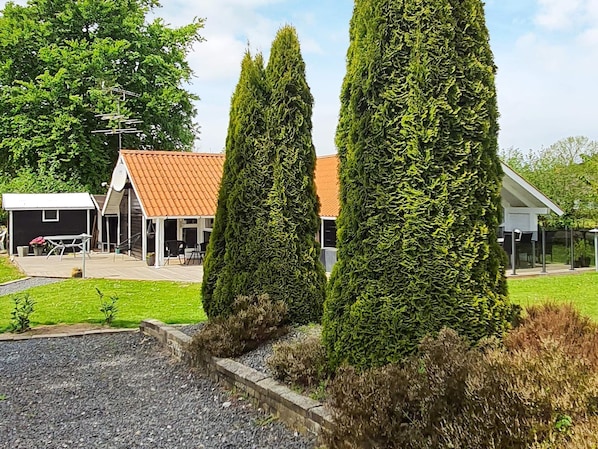 Un quartier résidentiel avec deux grands arbres à feuilles persistantes, bien entretenus, une maison avec un toit recouvert de tuiles rouges et un espace extérieur aménagé avec des sièges.