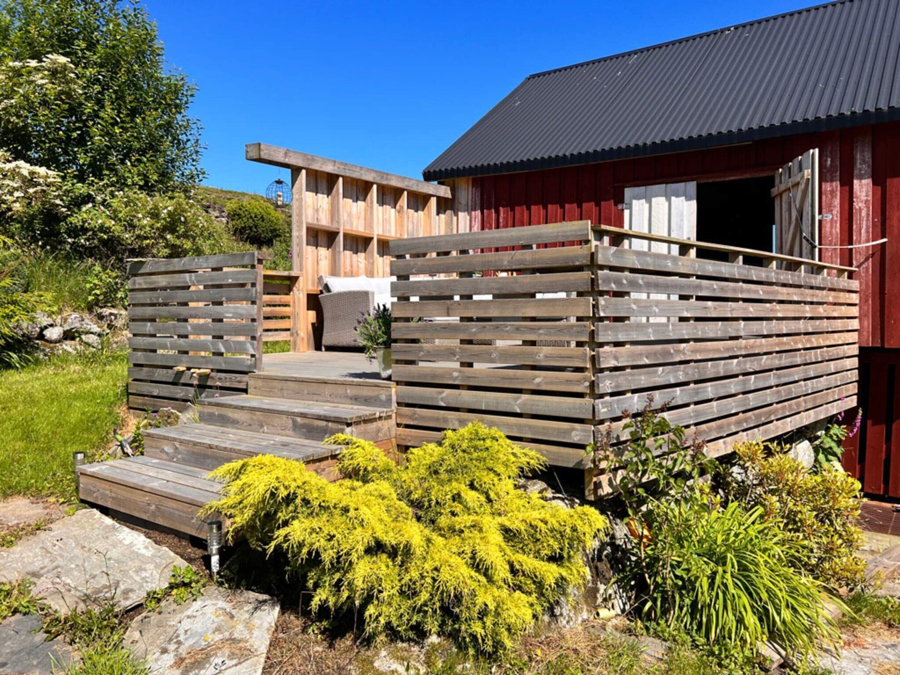 Una terraza de madera que conduce a un edificio rojo con techo de metal, rodeado de vegetación y un camino de piedra.
