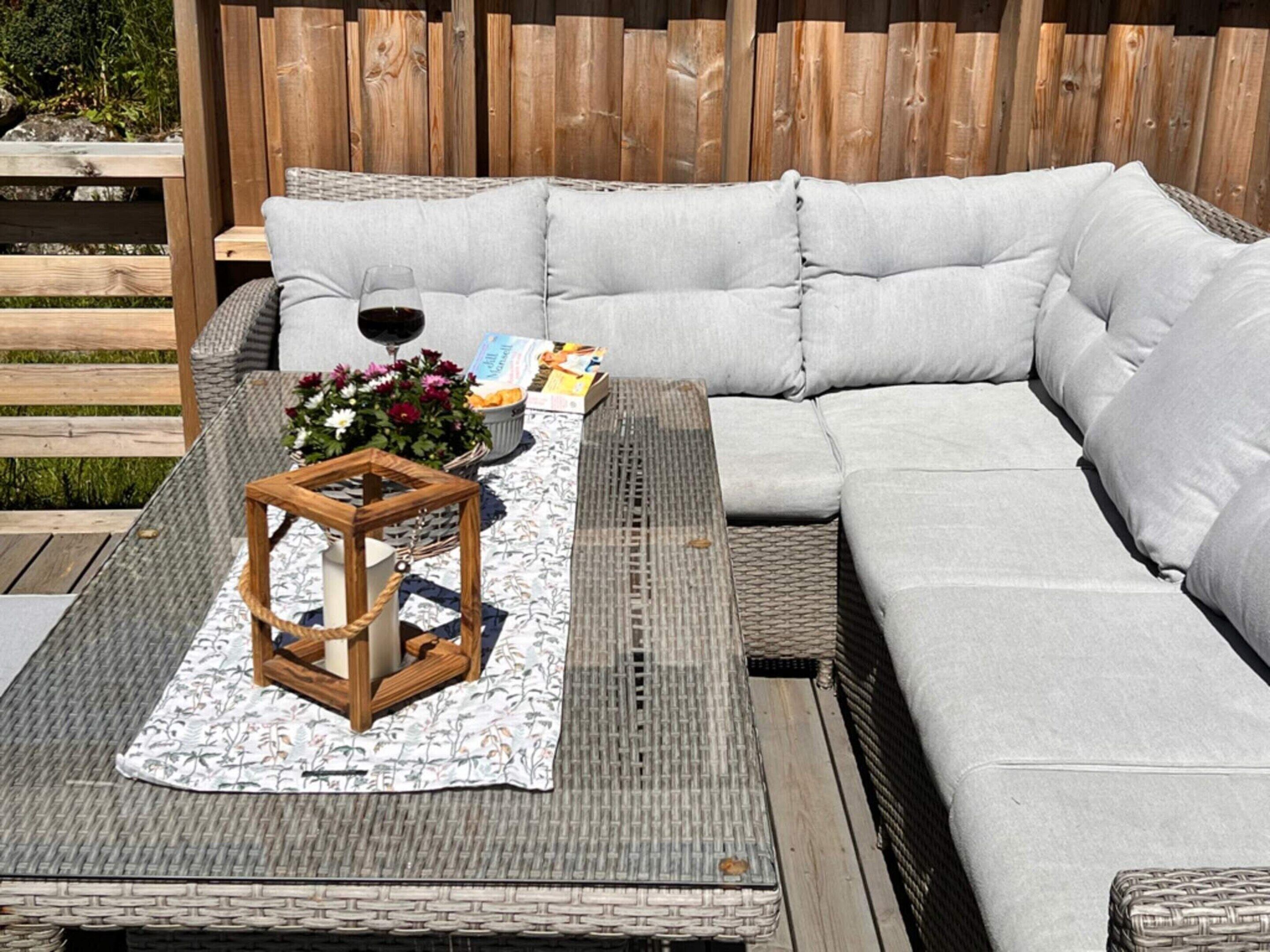 A wicker sofa with a grey cushion, a woven table with a wooden lantern, and a floral arrangement on a patterned table runner.