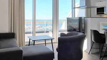 Ein modernes Wohnzimmer mit einer Couch, einem Sessel und einem kleinen Tisch. Ein Fernseher ist an der Wand befestigt, und durch große Fenster hat man Blick auf einen Yachthafen.