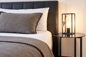 En seng med et mørkt sengegavl, hvide og grå puder, og et rundt, sort natbord med en lampe i vintage stil.