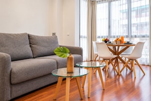 A modern living room with a grey sofa, a glass coffee table, and a dining area with a round table and white chairs.