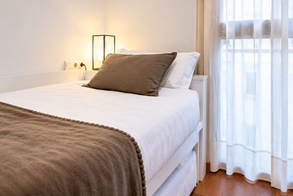 Una cama bien hecha con una manta y almohadas marrones, una lámpara de noche y cortinas translúcidas en una ventana.