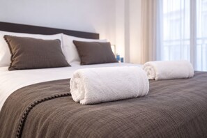 Cama bem arrumada com colcha marrom e toalhas brancas, cabeceira escura e janela com cortinas.