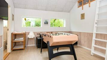Una habitación con mesa de fútbol, un estante blanco, una escalera y ventanas con vistas a plantas.