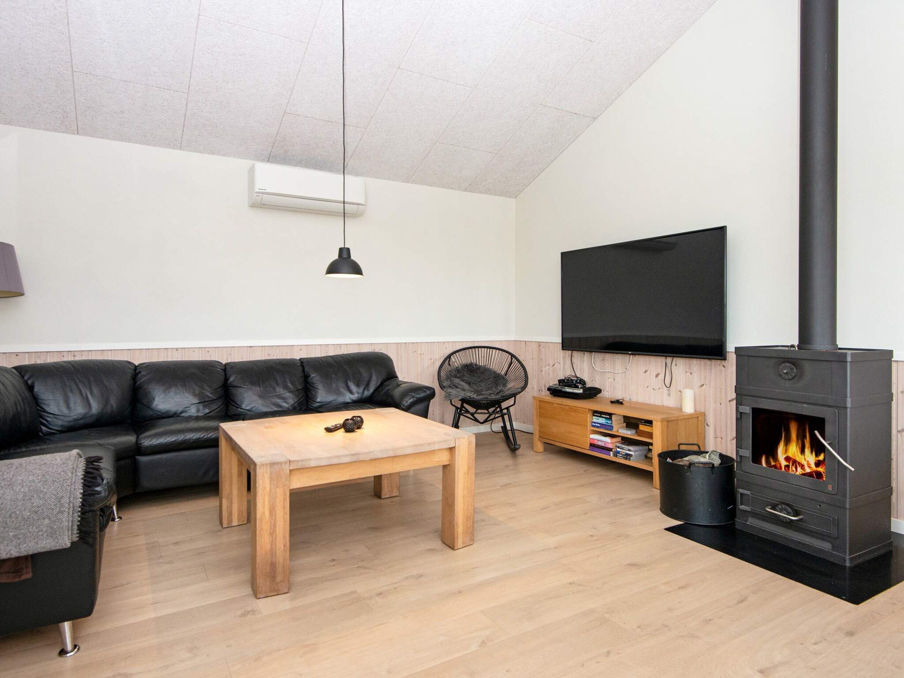 Uma sala de estar com sofá de couro preto, mesa de centro de madeira, ventoinha preta, televisão e lareira a lenha.