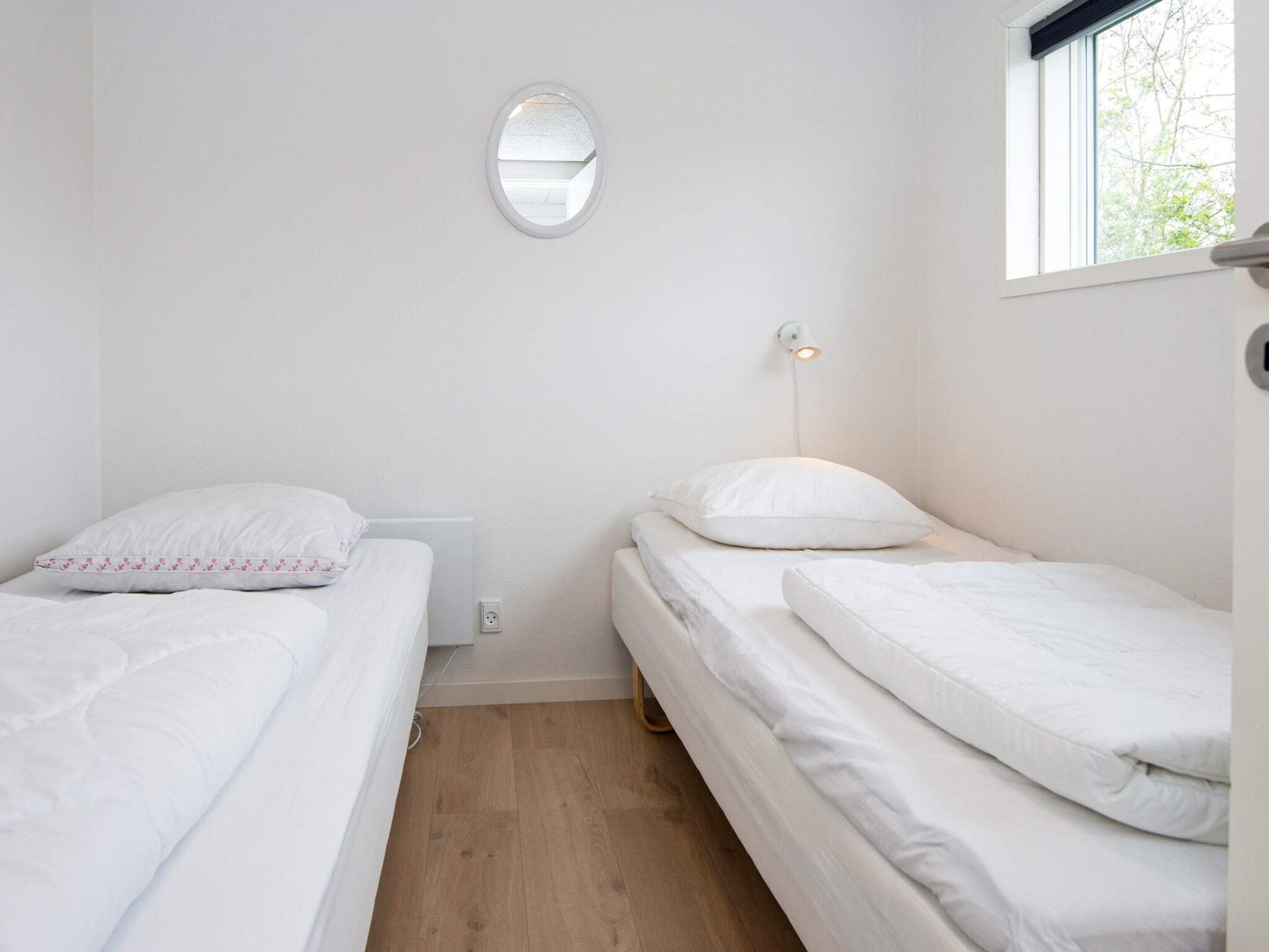 Un alojamiento pequeño, con una sola habitación, dos camas, una mesita de noche, un espejo en la pared y un ventanal que proporciona luz natural.