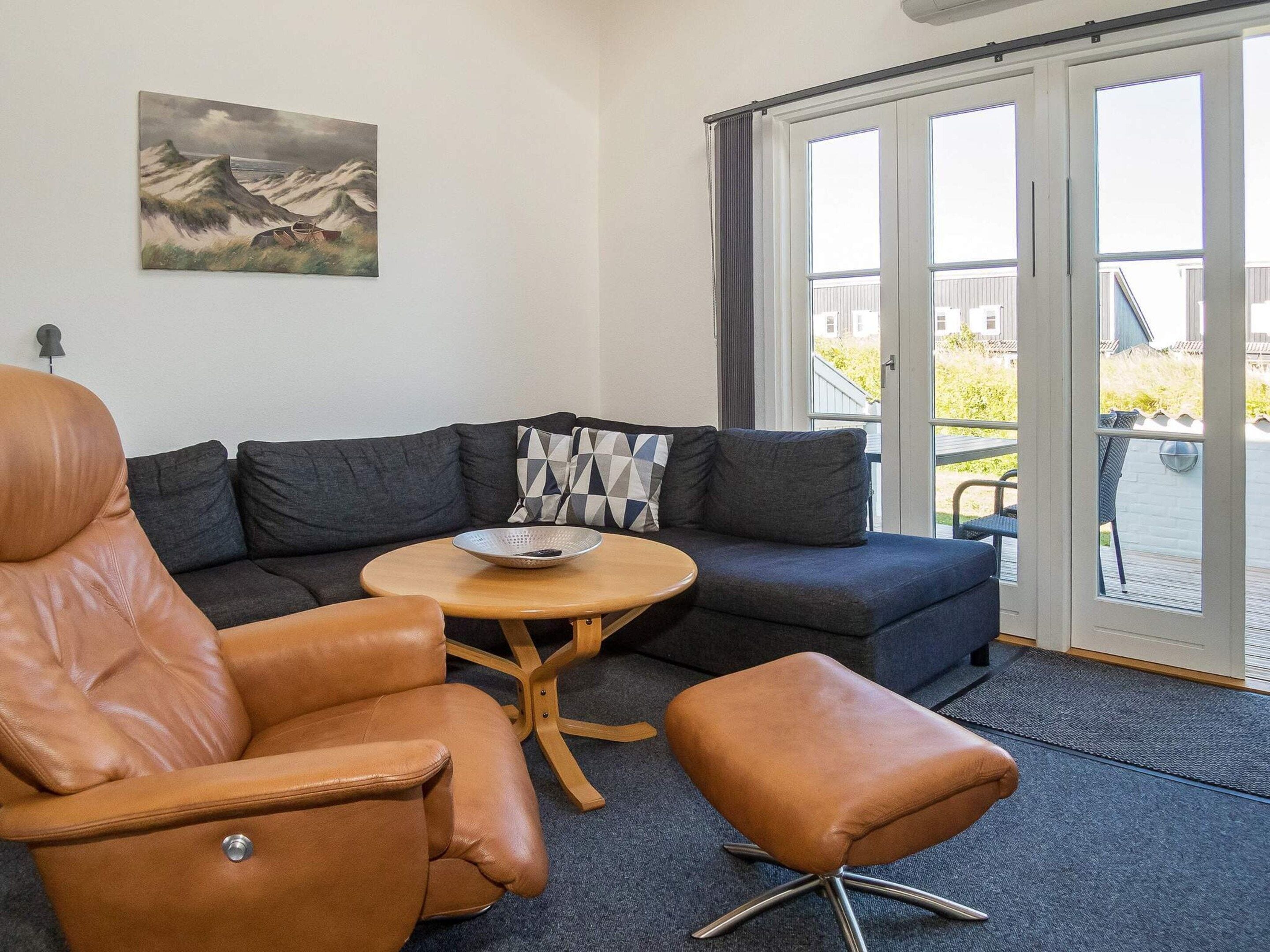 Un salon moderne avec un fauteuil en cuir, un canapé et un pouf. Un tableau orne le mur et on peut admirer le paysage à travers les portes vitrées.