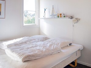 Habitación individual con una cama y edredón blancos, ventana con vista a árboles y repisa con varios objetos, incluyendo un barco modelo.