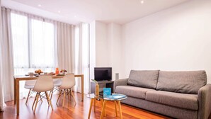 A modern living room with a grey sofa, wooden dining table, and chairs. A flat-screen TV is on a stand, and a small table with a glass top is in the center. - Sweett | Eixample 2D (Barcelona)