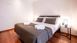 A neatly made bed with a brown bedspread and white linens, a bedside table with a lamp, and a wall socket. - Sweett | Eixample 3D (Barcelona)
