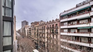 A row of multi-story residential buildings with balconies, a narrow street with parked vehicles, and a tall building in the background. - Sweett | Eixample 3D (Barcelona)