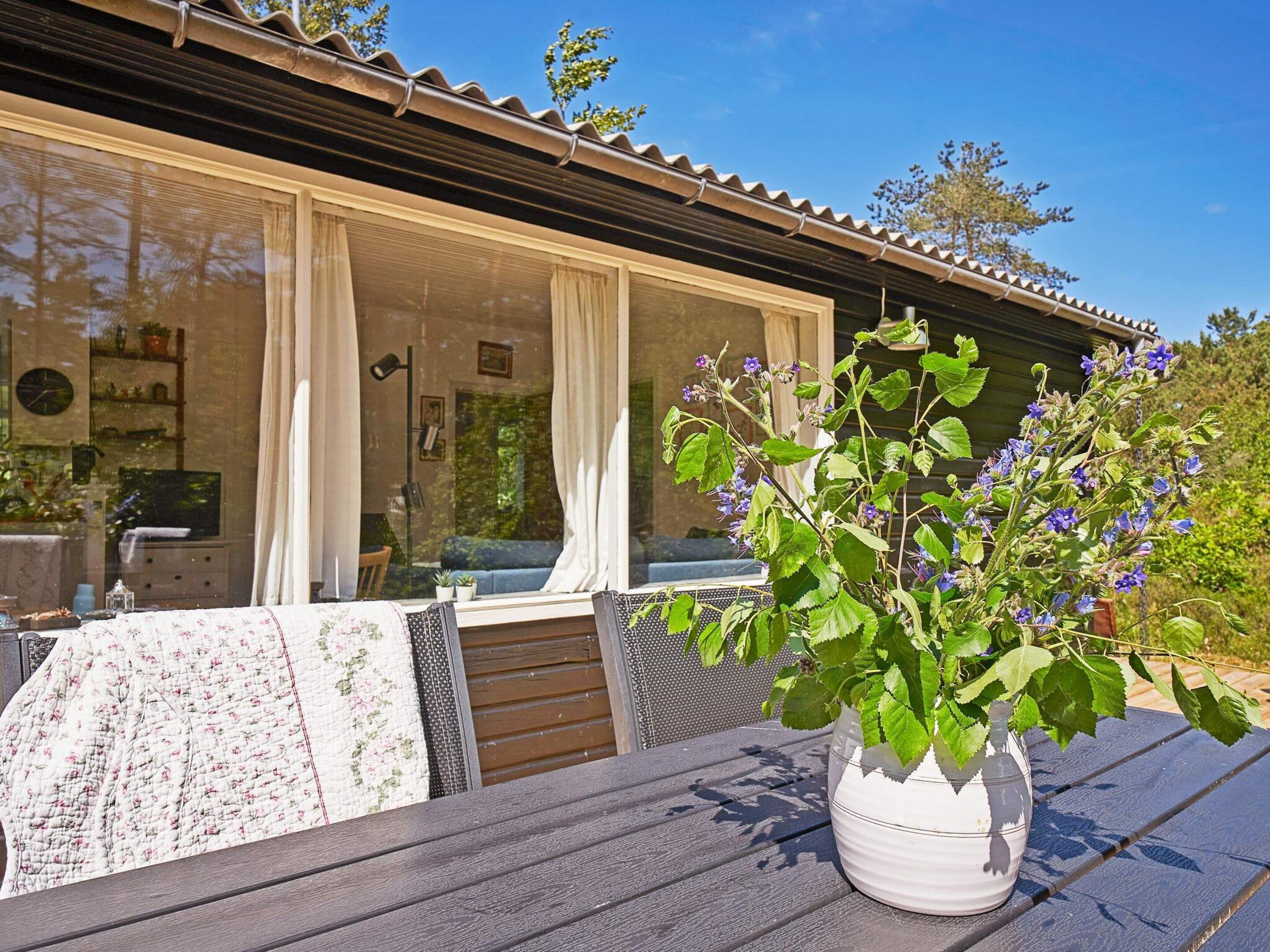 Un espacio al aire libre acogedor con una mesa, una planta en maceta y vistas a una casa con amplias ventanas y cortinas.