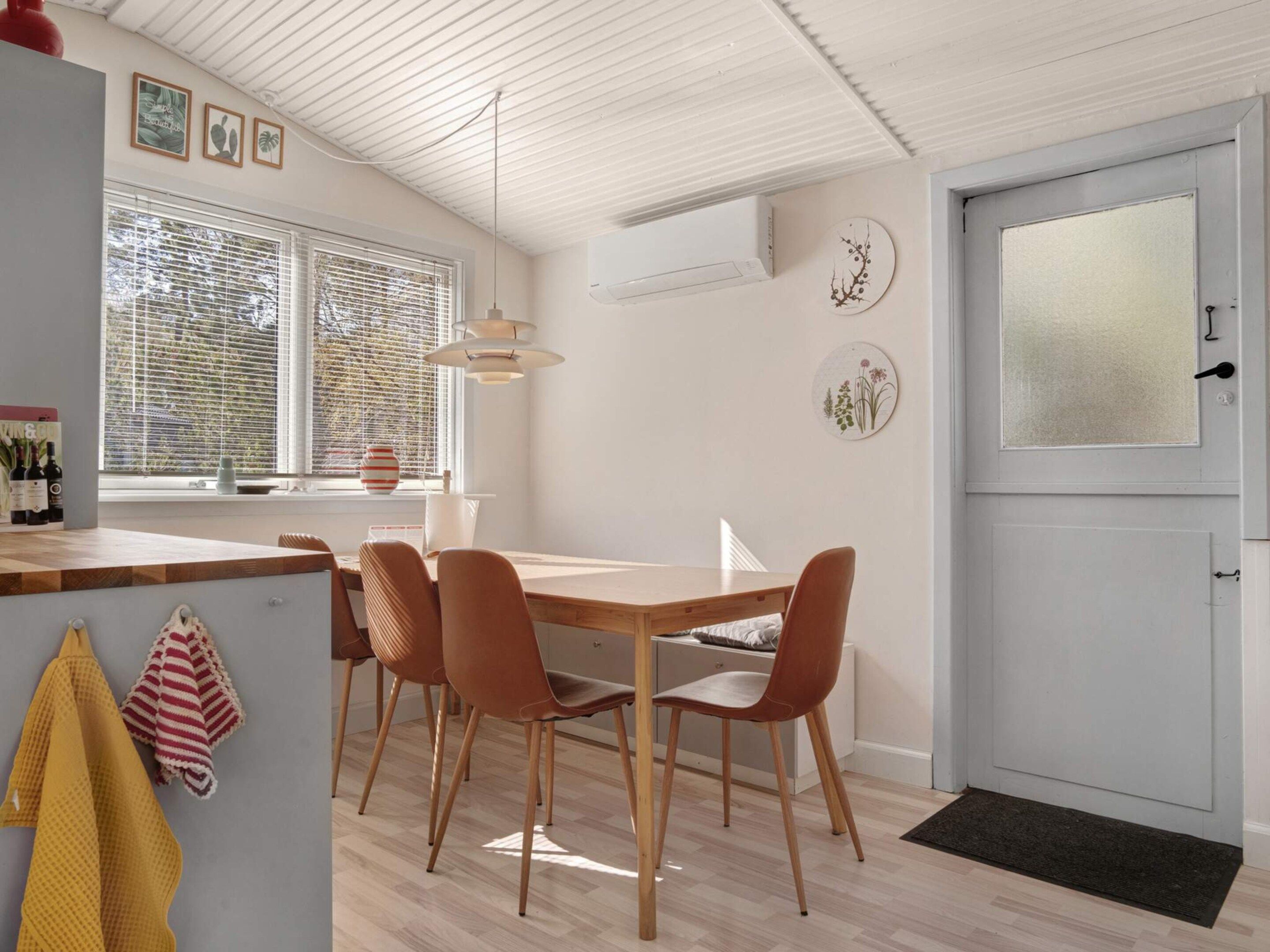 A modern kitchen with a dining area, featuring a wooden table and chairs, a window with blinds, and a door with a frosted glass panel.